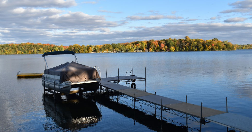 A covered pontoon boat sits on a lift beside a dock on a calm lake, with colorful autumn trees along the shore.