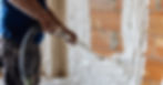 A man in a blue shirt is using a spray foam hose to cover a wall in foam. The exposed parts of the wall are orange.