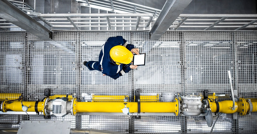 An individual in a blue uniform and yellow hard hat using a tablet while walking next to yellow pipes.