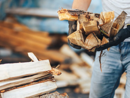 A person wearing gloves holds a pile of split firewood, with more firewood stacked on the ground in a blurred background.