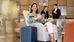 A woman sits in a hotel lobby chair. She holds a smartphone as she sits beside a blue suitcase near a reception desk.