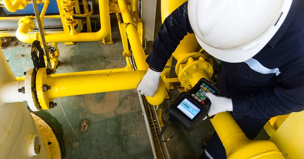 A worker in a white hard hat checks bright yellow industrial pipes with a handheld ultrasonic meter.