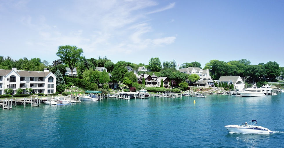 A recreational boat cruises across a clear blue lake past waterfront homes and tree-lined shores on a sunny summer day.