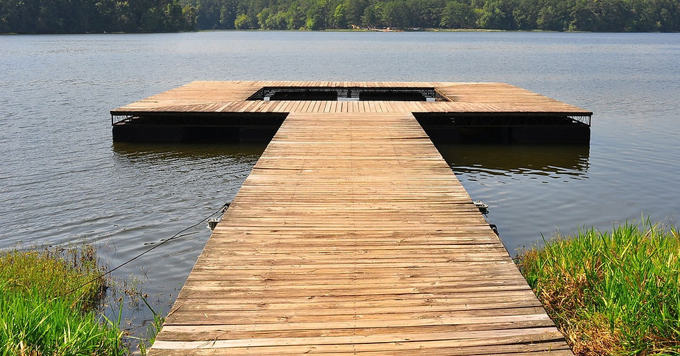 A large wooden dock floats on a blue lake, with green trees lining the shore under a bright, sunny sky.