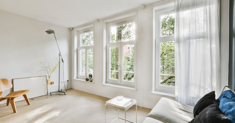A sunlit living room with a white sofa, round wicker coffee table, hanging plants, and large windows overlooking trees.