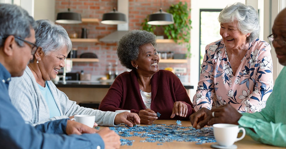 A group of seniors sit at a table together. They smile and work on a blue puzzle while also drinking from coffee cups.