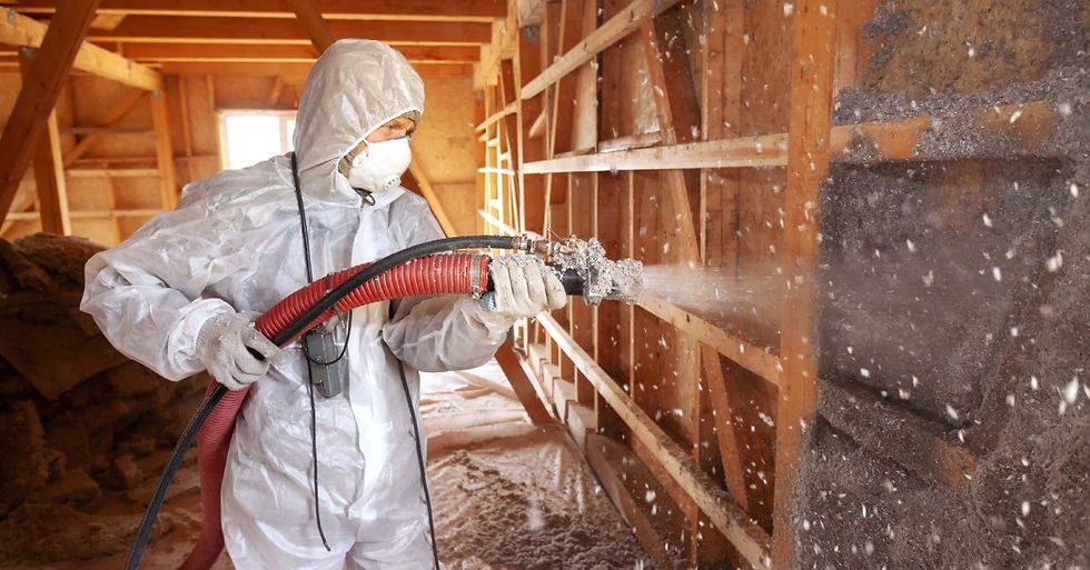 A person wearing personal protective equipment, including gloves and a mask, holding a hose and spraying cellulose into a wall.