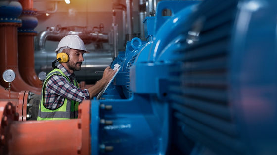 A man wearing a white hard hat and a bright yellow safety vest writes on a clipboard, standing next to an industrial pump.