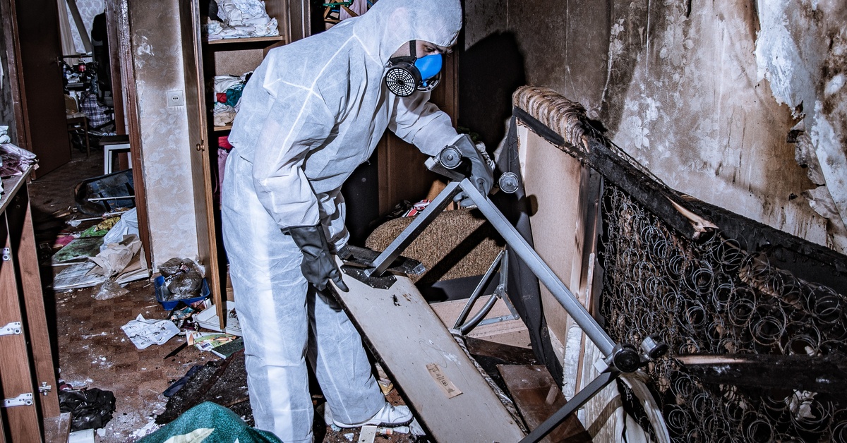 A person in a protective suit is picking up items in a burnt down structure. He is wearing a blue breathing mask.