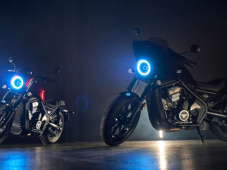 Two motorcycles parked on a polished concrete floor inside a room with dim lights, with both headlights on.
