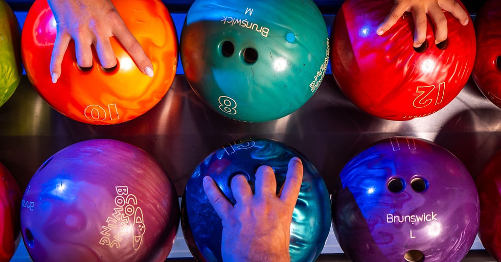 Several colorful bowling balls rest in the ball return. There are three hands, each grabbing a different ball.