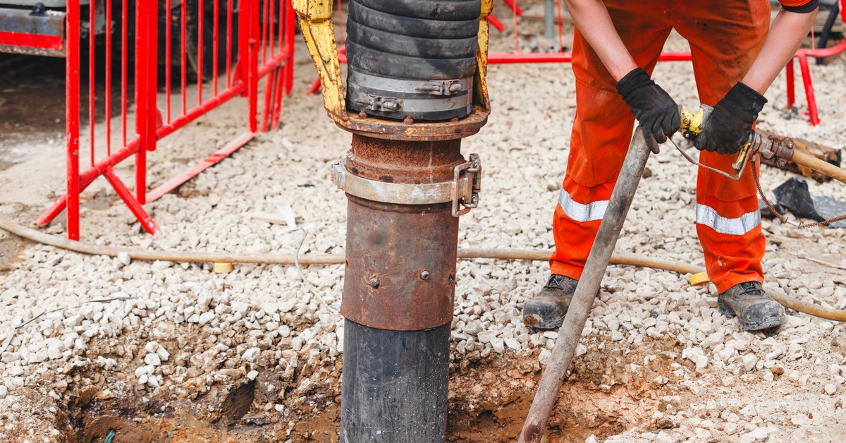 A man in a red jumpsuit is bending over while he is holding a hose. There is also a metal pipe going down into a hole.