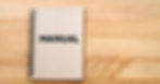A brown spiral notebook with the word "MANUAL" in black font on the cover rests on a wooden surface.