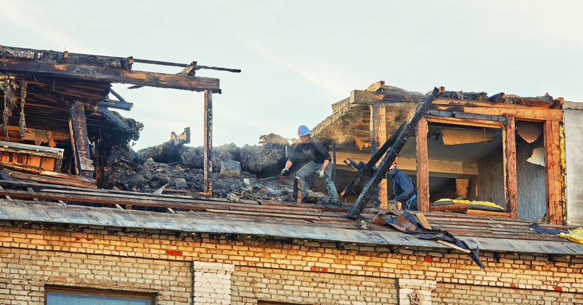 A building that has sustained fire damage on its room has several people on top inspecting the damage.