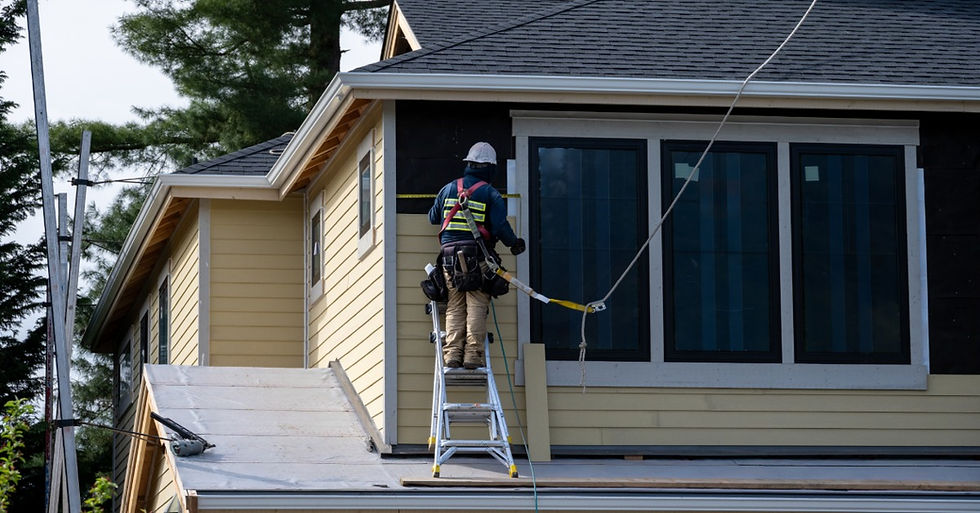 A carpenter wearing a hard hat and harness standing on a ladder while installing wood siding on a house exterior.