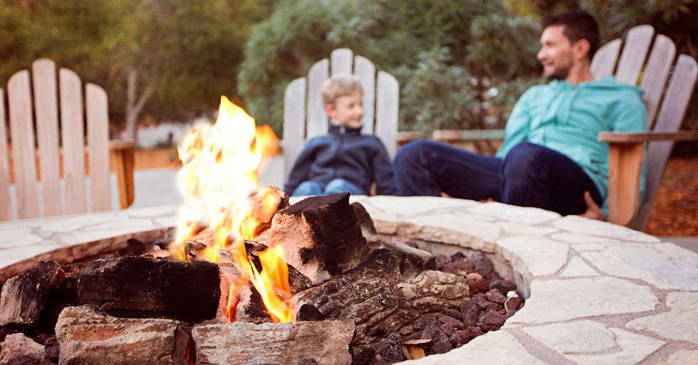 A man sitting on a chair outside beside a child as they talk to one another. There's a firepit in front of them and a forest in the background.