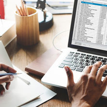 A woman sitting at a table in front of her computer going through documents while using a spreadsheet program.