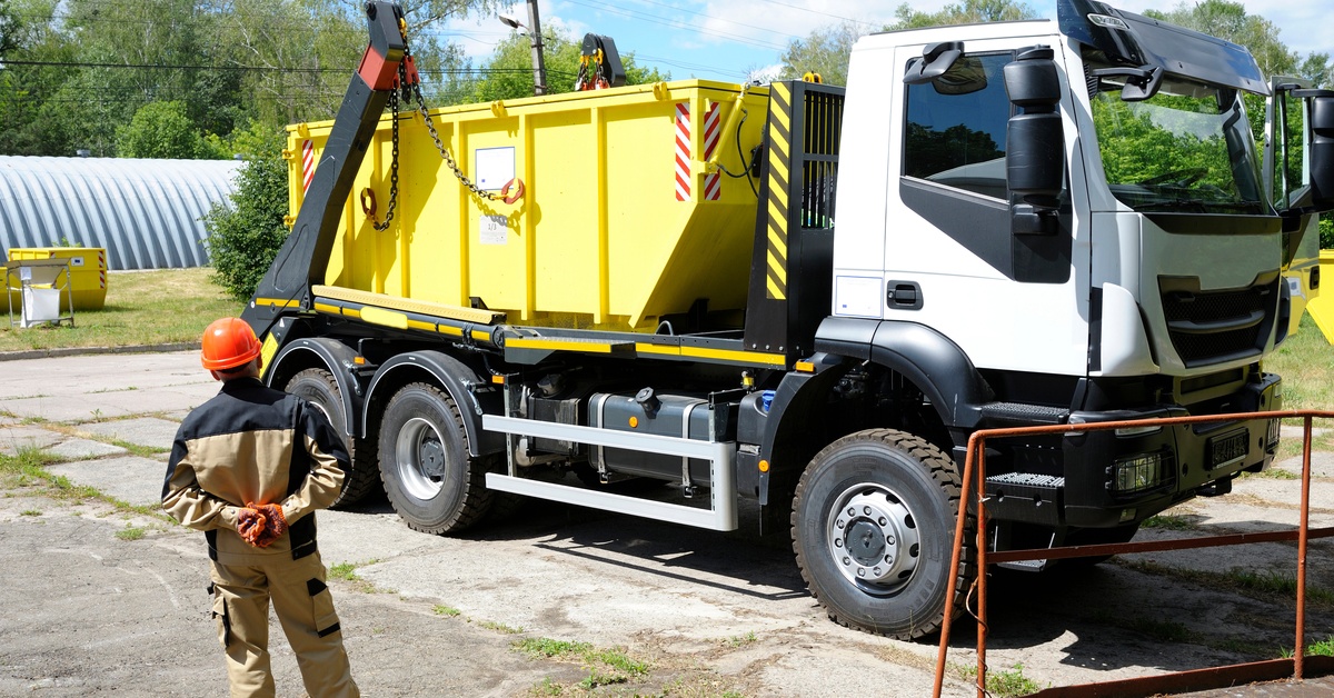 A truck with a yellow container on its bed is parked on a road. A man in protective gear is standing next to the truck.