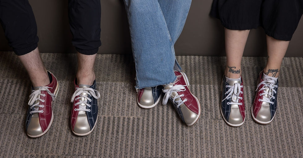 Three pairs of legs wearing bowling shoes. The person in the middle has their legs crossed at the ankle.