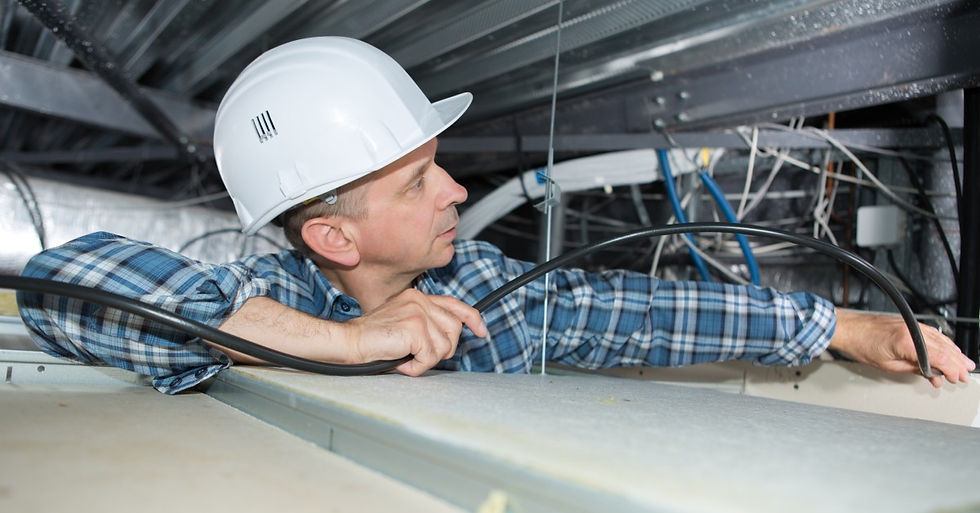 A man in a white hard hat and a blue plaid shirt is looking at some wires that are hanging from a ceiling.