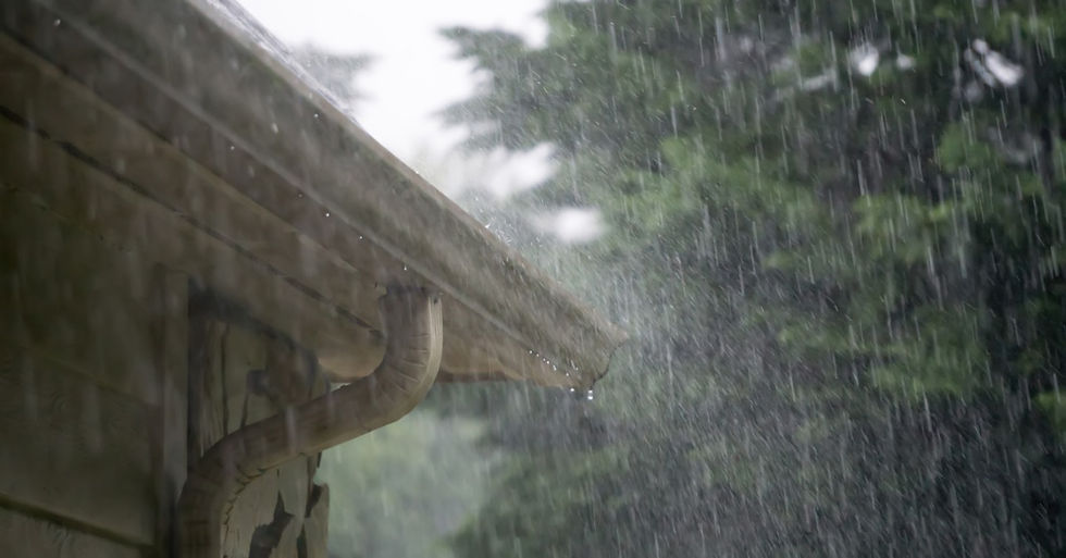 The edge of a house's roof with heavy rain pouring down over it. There are tall trees behind the house.