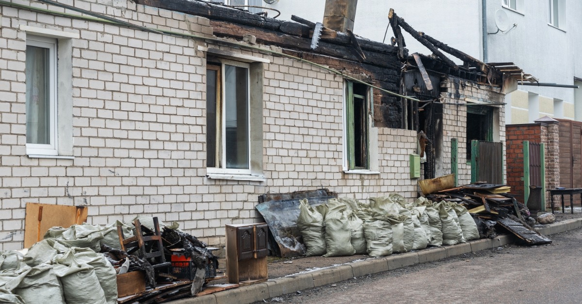 A building with white brick siding has signs of fire damage along its roof. There are garbage bags lined up on the curb.
