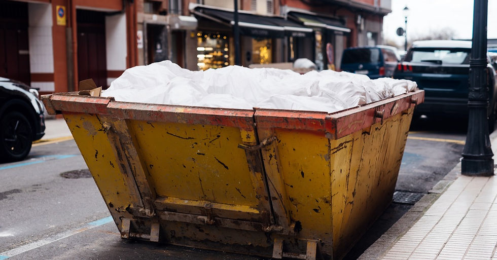 A large dumpster is set up on a street; there is a car parked next to it. In the background are buildings.