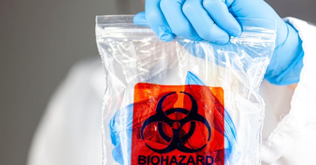 A person wearing blue gloves and a white jumpsuit is holding a bag with a biohazard warning on it.