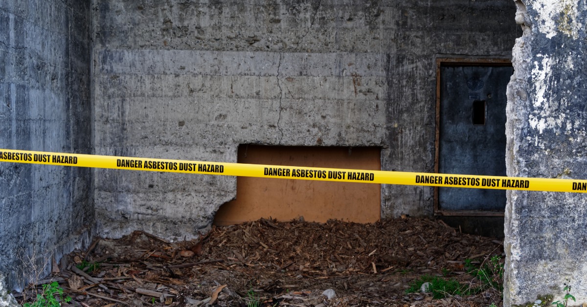 A dilapidated room is taped off with a yellow strip that warns of asbestos exposure. There is dirt on the ground.