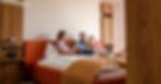 A family of four sits together on a large bed in a hotel room and looks through a booklet of information.