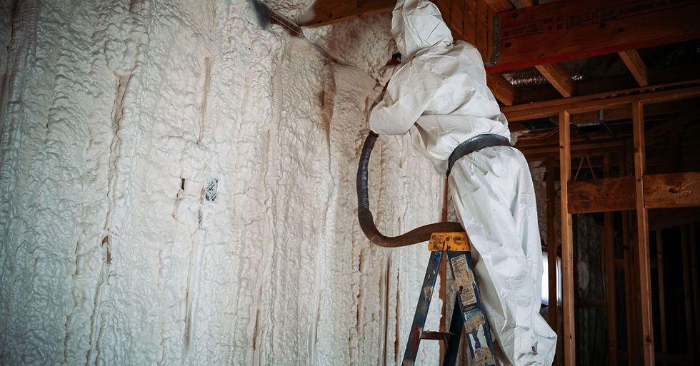 A person wearing white coveralls applies spray foam insulation to the interior spaces of a wall while standing on a ladder.