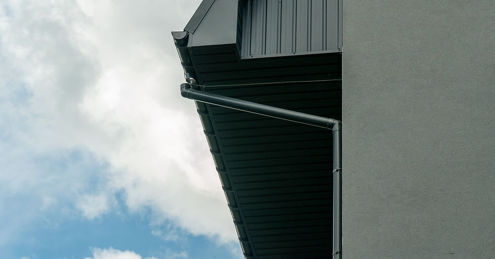 A commercial building corner with a dark metal gutter and downspout beneath a roof overhang on a cloudy day.