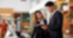 Two hotel employees in black suits stand behind the reception desk and look at something on a tablet.