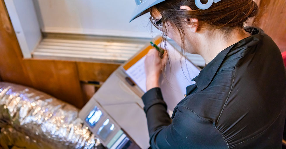 A woman in a hard hat and safety glasses taking notes on a clipboard while standing in front of a silver air duct.
