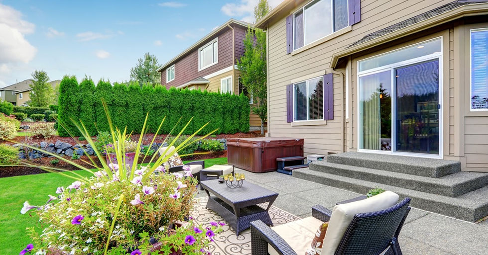 A backyard patio with wicker chairs, a table, a hot tub, steps, a sliding glass door, flowers, a lawn, and shrubs.