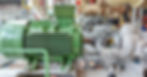 A green electric motor with metal vents and mounting bolts sits in front of industrial pipe assemblies.