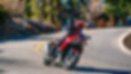 A motorcycle rider on a red-and-black adventure motorcycle turning on a paved road in a wooded area.