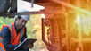 A man in a helmet and reflective suit writing on a clipboard while inspecting an electric brake system beneath a railcar chassis.