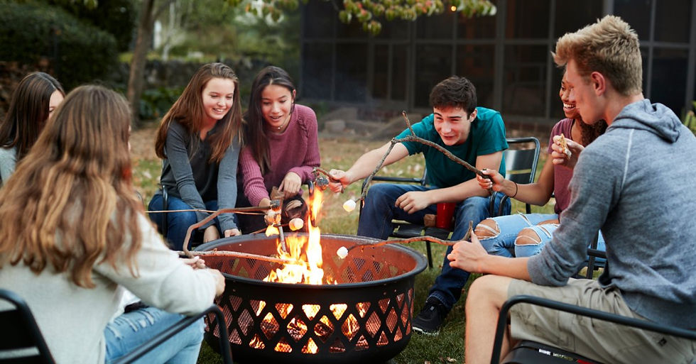 A group of seven friends sitting outside, around a bonfire, and roasting marshmallows on sticks over the flames.
