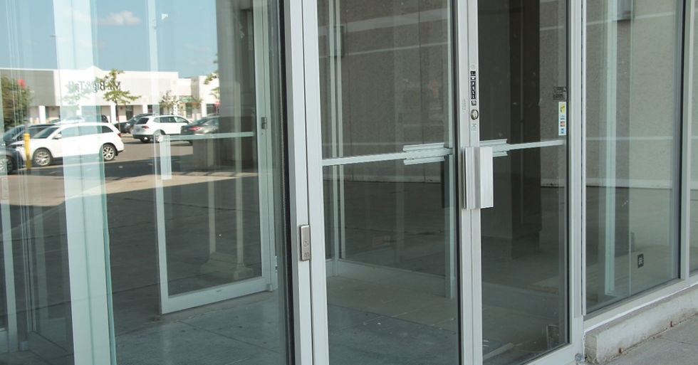 A glass storefront entrance features double doors that reflect parked cars and the sky. The space inside appears empty.