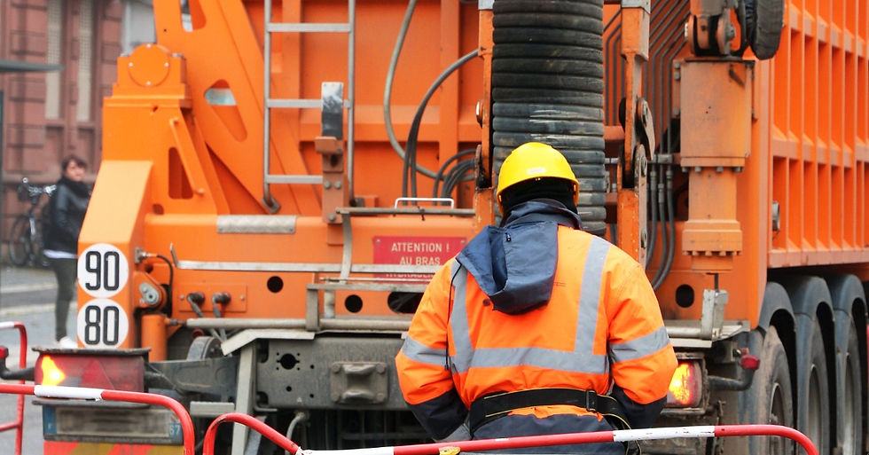 A man dressed in orange I standing next to an orange truck. The truck has a black hose coming out of it.
