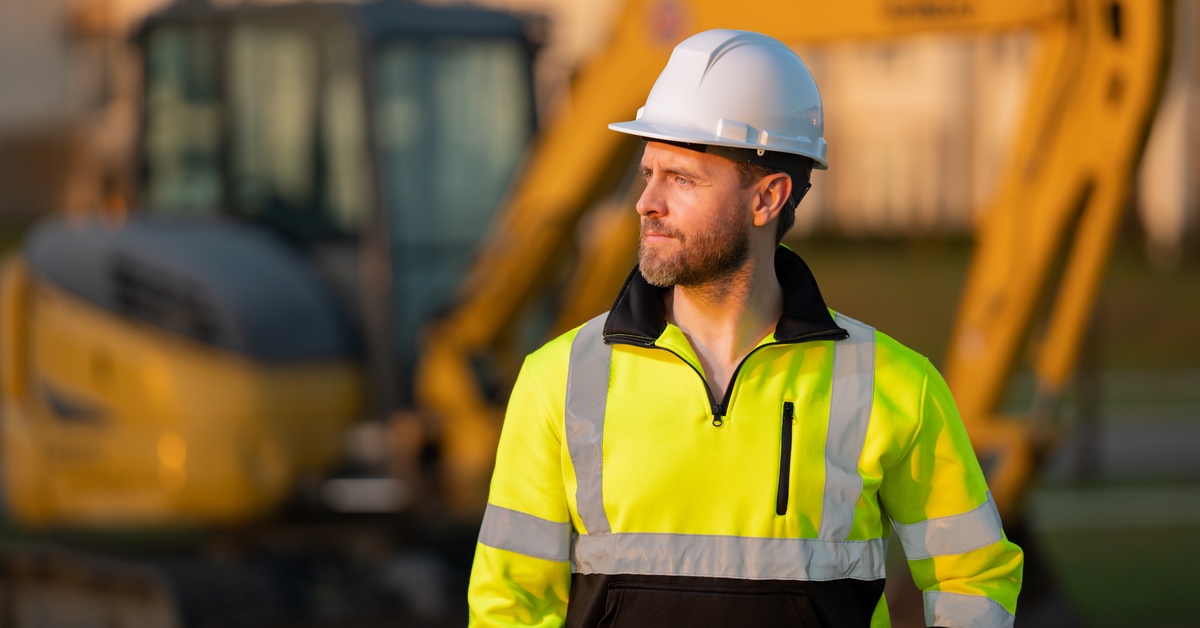 A man in a high-visibility jacket and a white hard hat is walking away from construction equipment.