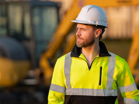 A man in a high-visibility jacket and a white hard hat is walking away from construction equipment.