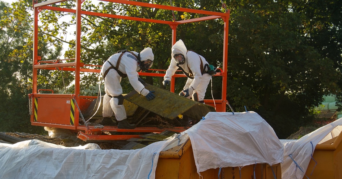 Two people in protective gear are holding a board and are throwing it into a dumpster.