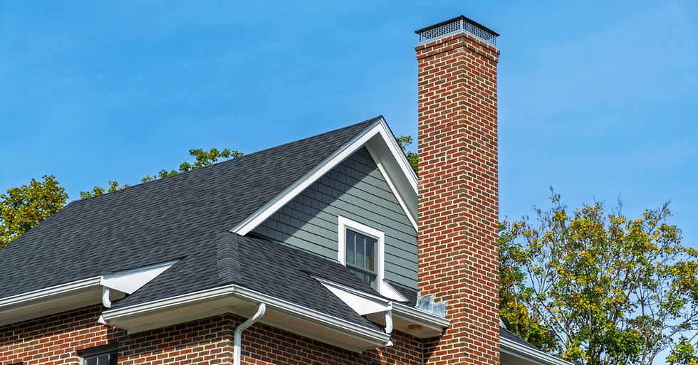A brick home with a tall chimney cap rising above a dark shingled roof, framed by trees beneath a clear blue sky.