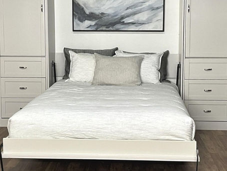 A white wall bed with side cabinets, drawers, gray-and-white pillows, framed art, a potted plant, and wood flooring.