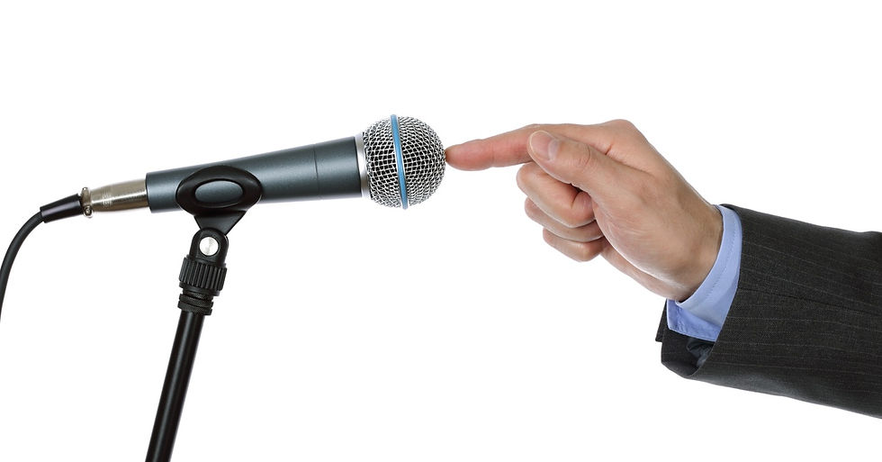 A person in a suit jacket and blue shirt taps a microphone on a stand with their finger against a white background.