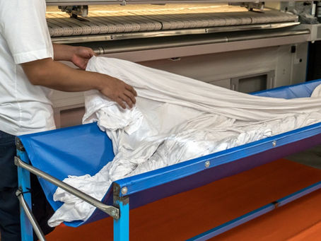 Two workers standing on either side of a bin with laundry in it while placing a clean sheet into a folding press.