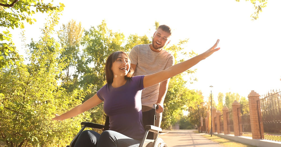 A young man guides a young woman in a wheelchair through a path in a park as they enjoy the sunny day.