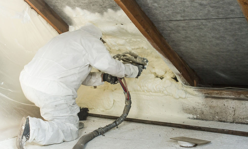 A worker wearing a full-coverage bodysuit kneels over while holding a spray nozzle and hose to apply insulation under a roof.